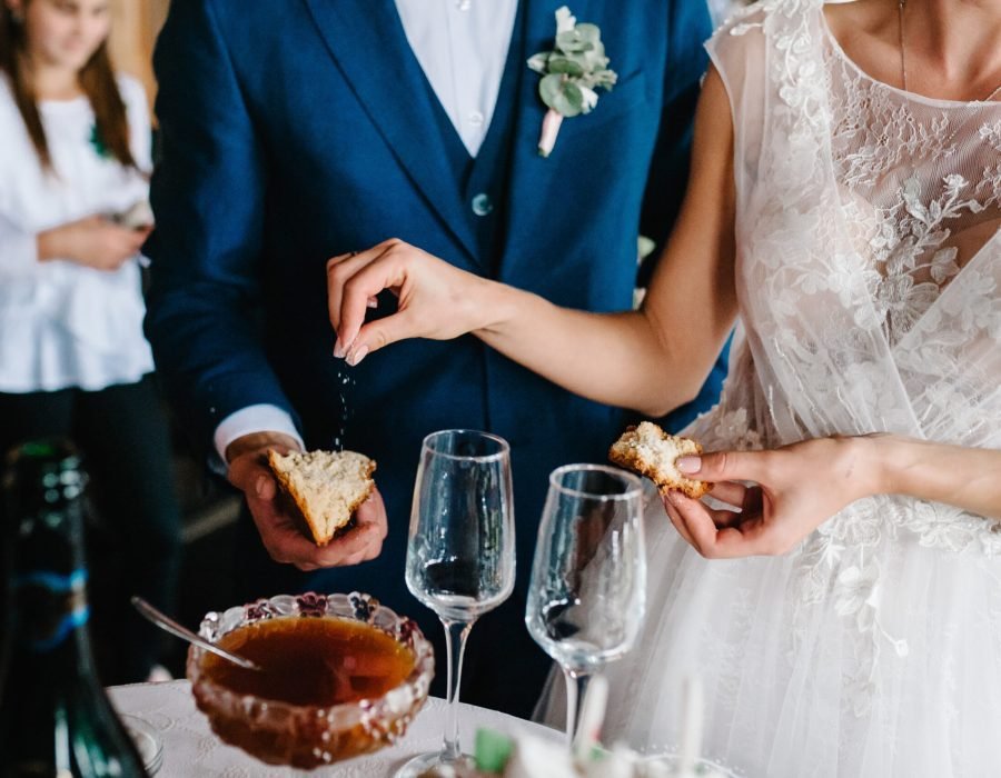 Close Up photo of bride and groom eating traditional loaf. Ukrainian and Russian wedding ceremony: bread with salt. Honey and glasses of champagne on an old rustic wooden table. Wedding traditions.